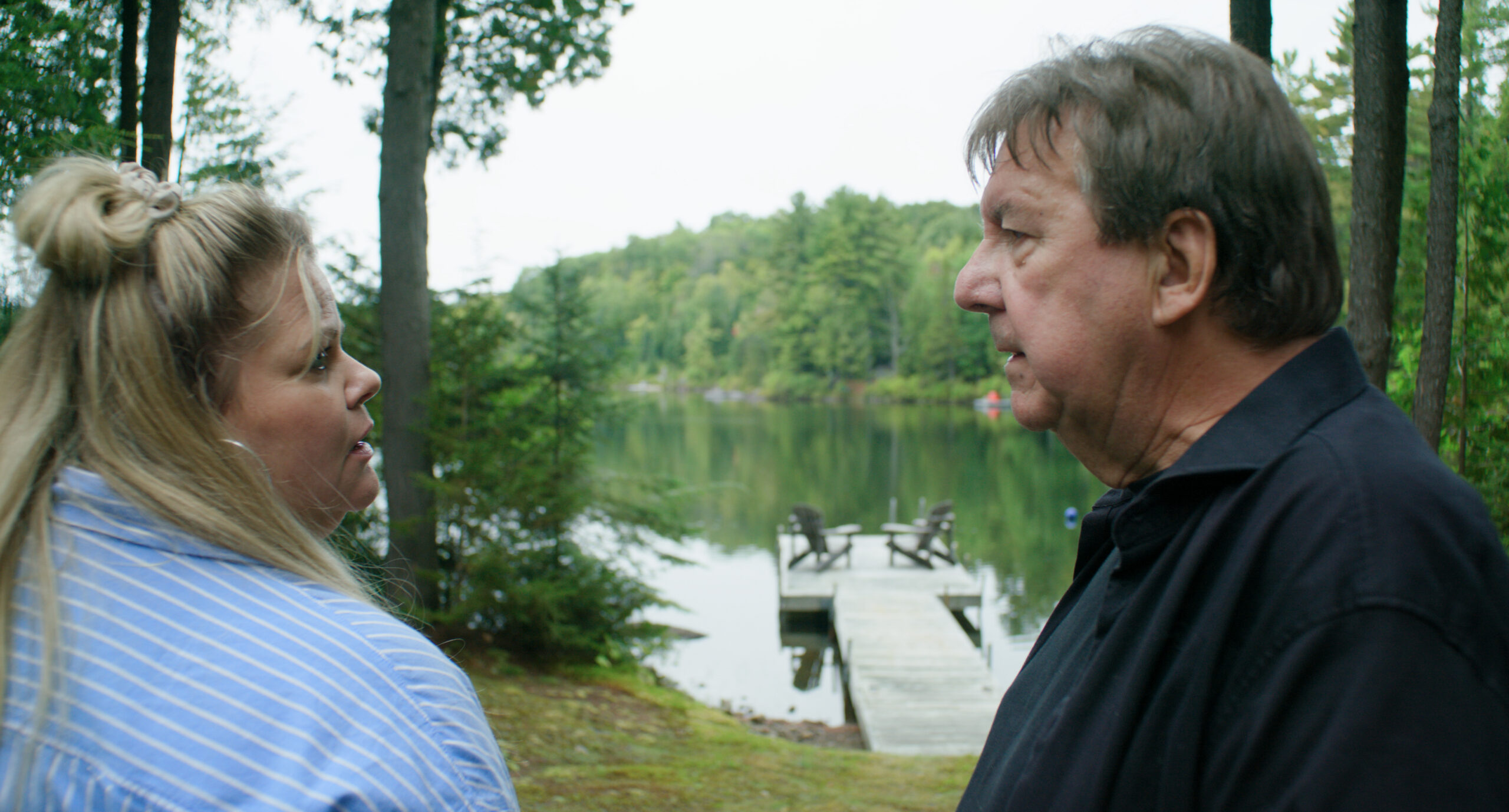 A blonde woman in a striped shirt and an older man in a dark shirt talk beside a lake with a wooden jetty, in 125, rue des Malaises