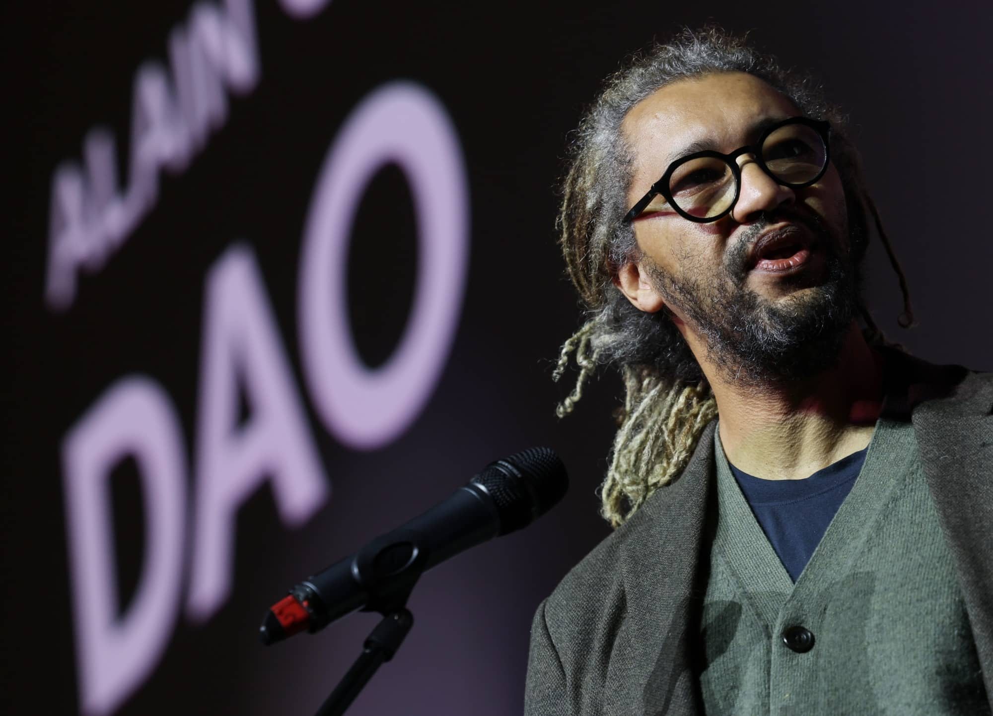 Alain Gomis, in round black glasses, speaks at a microphone in front of a screen showing the title Dao