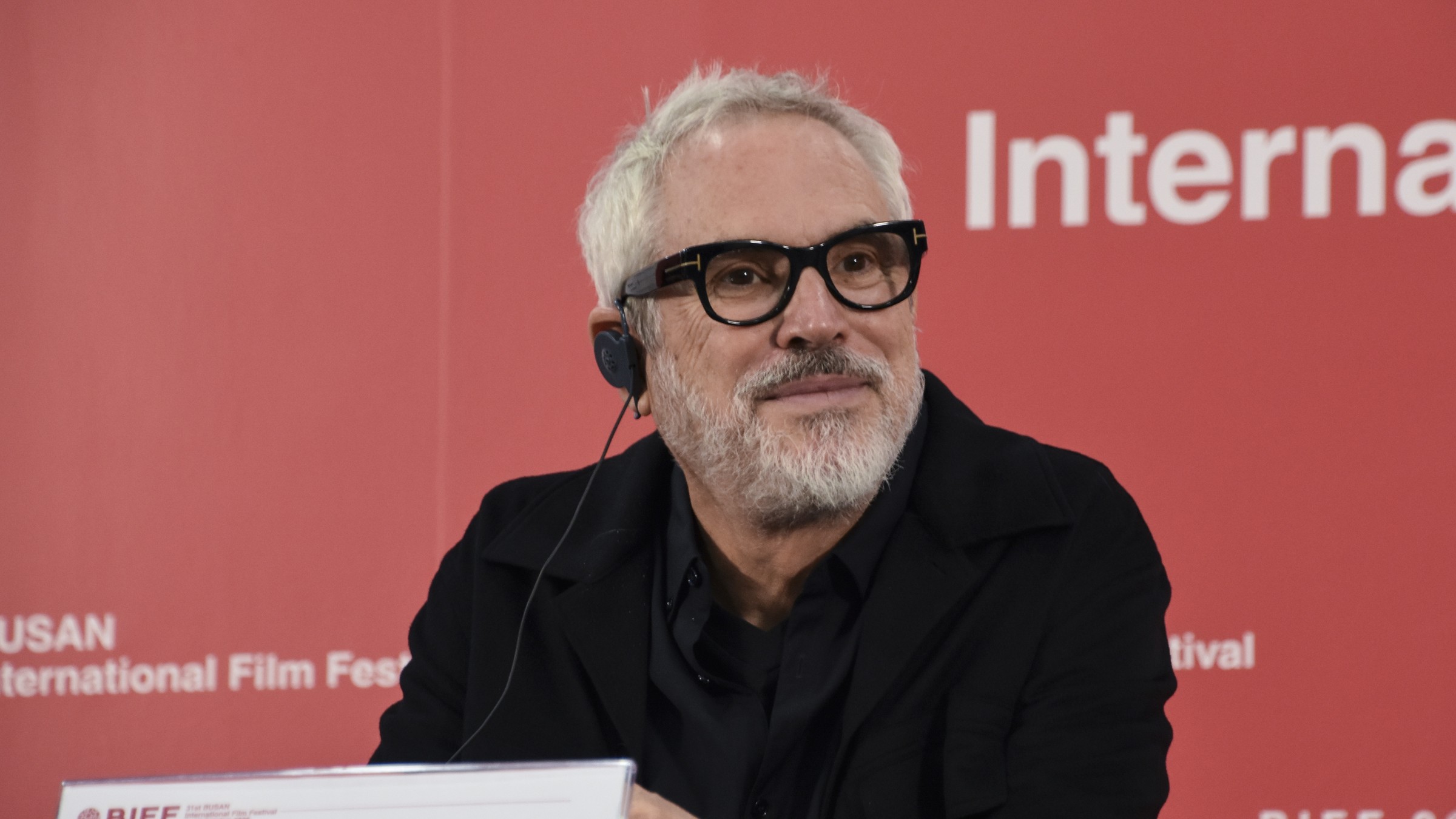 Alfonso Cuarón, grey-haired with a white beard and thick black glasses, smiles at a table in front of a red Busan festival backdrop