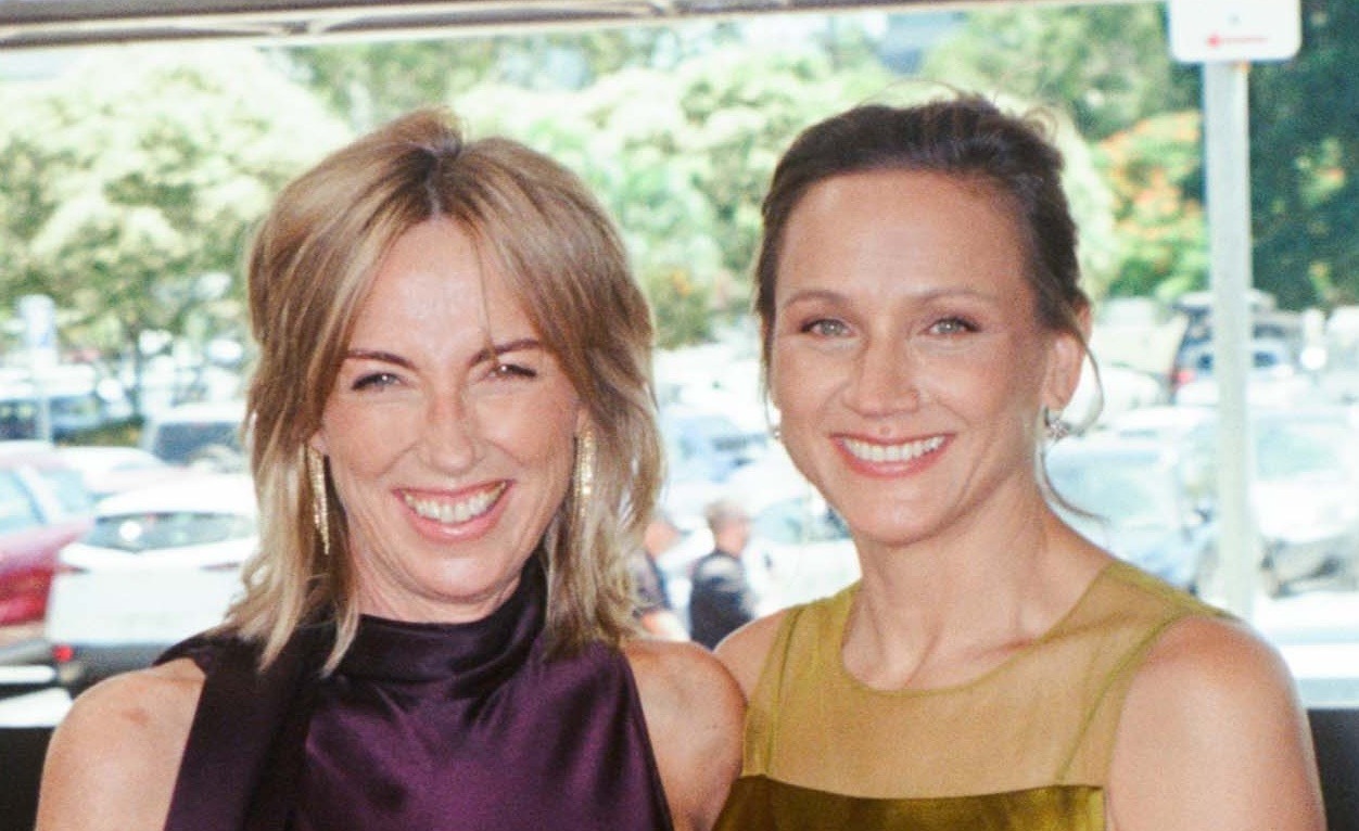 Two smiling women stand side by side outdoors, one in a dark purple halter top and one in an olive green top