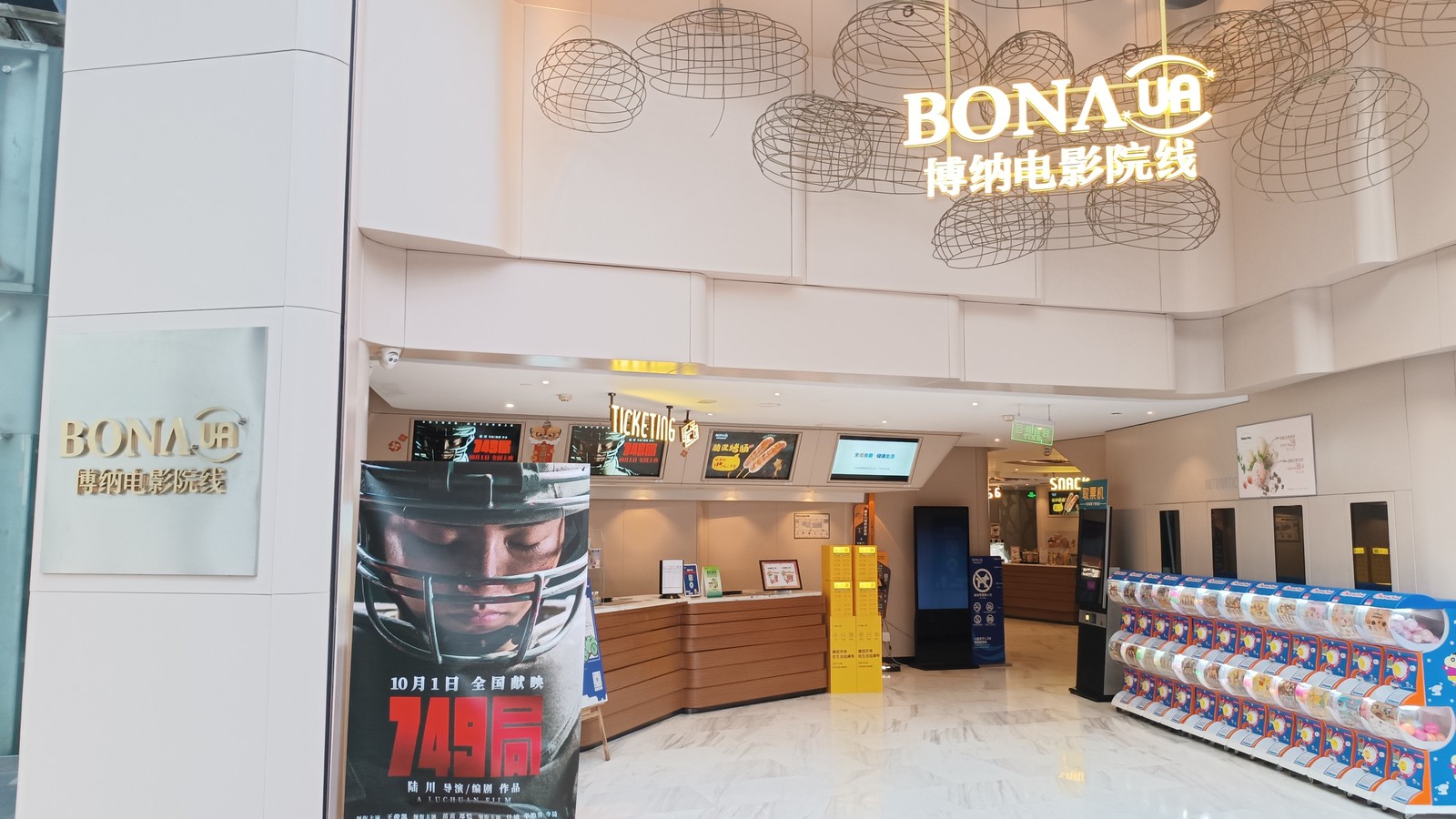 The ticket counter of a Bona UA cinema in a shopping mall, with an illuminated Bona UA sign overhead and a film poster standing at the entrance