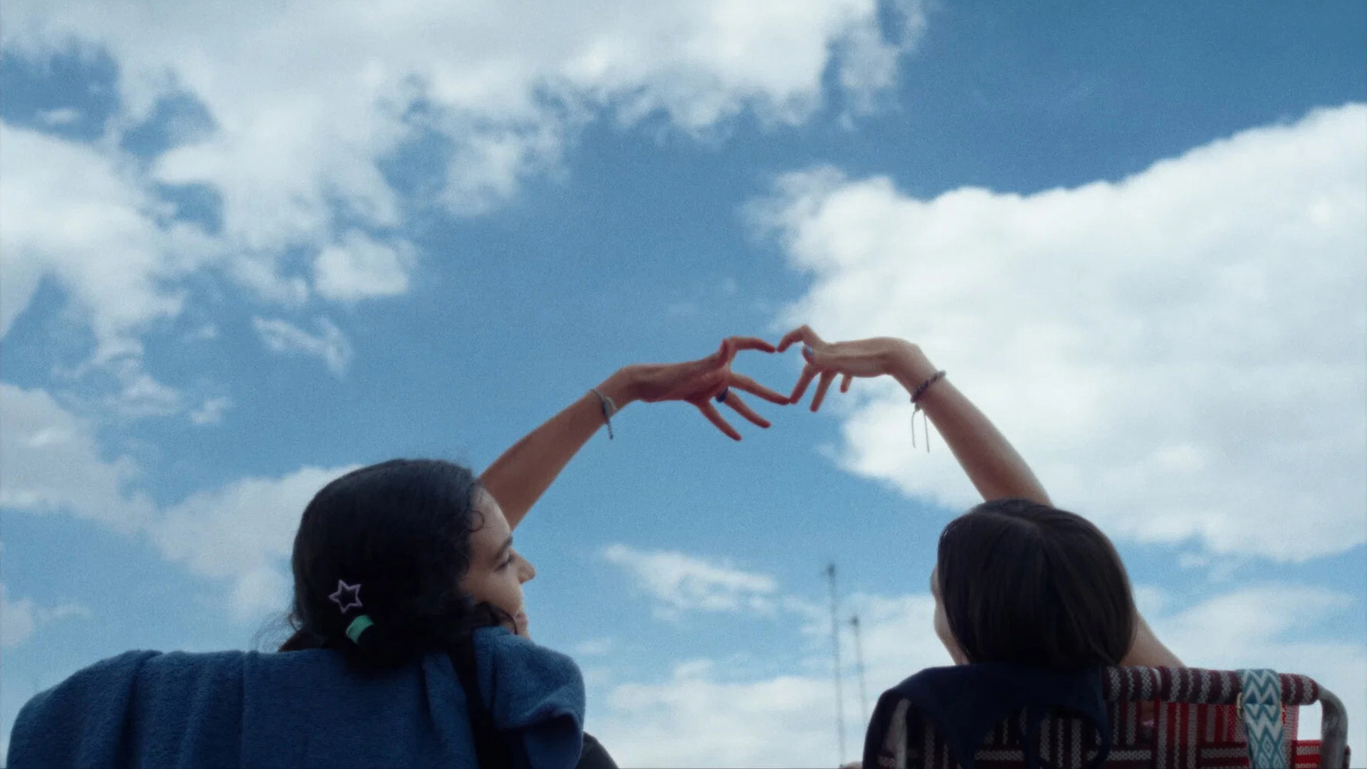 Two teenage girls seen from behind, raising their hands to form a heart against a blue sky, in Sad Girlz