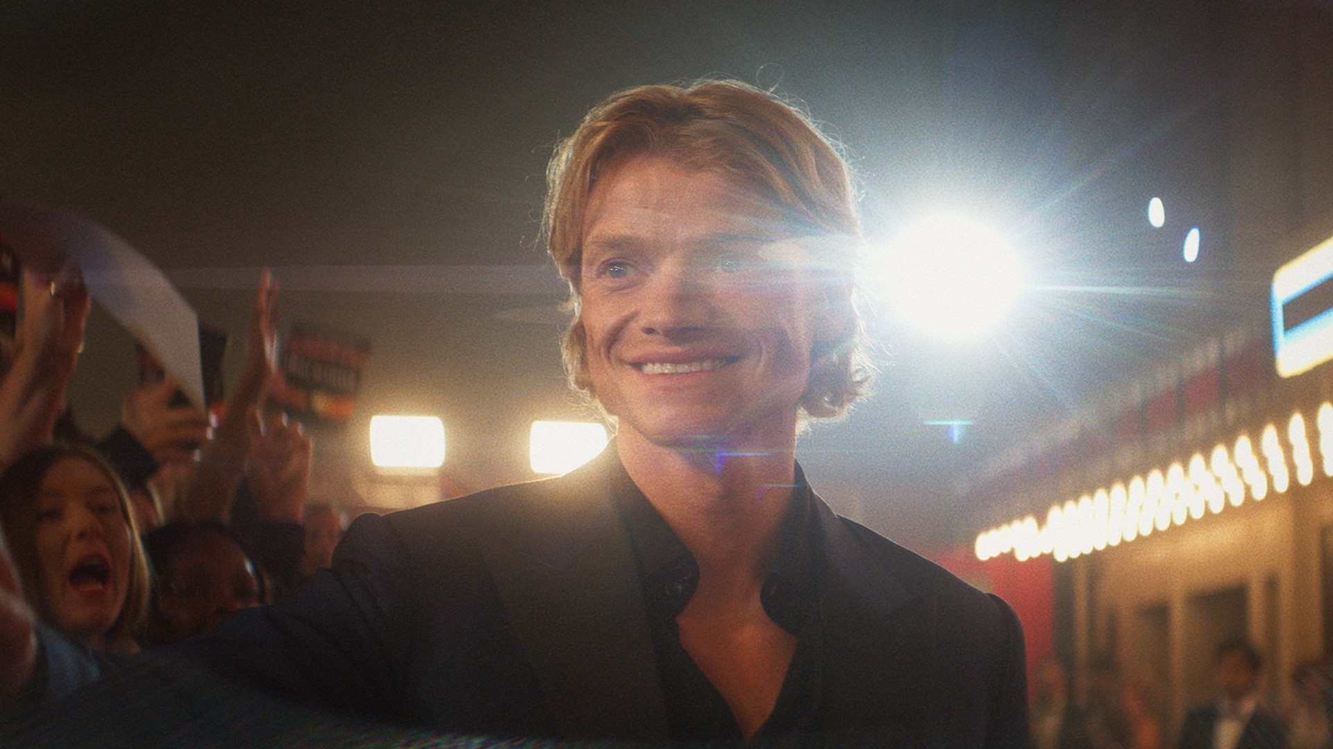 A man smiling on a red carpet as fans cheer behind him, in a still from Clayface