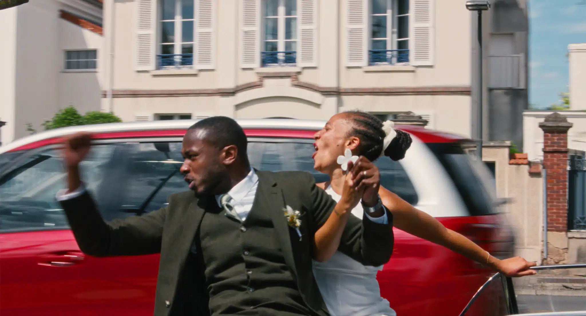A groom in a dark green suit and a bride in white with a flower in her braided hair lean out of a moving car, arms raised, on a sunny street of pale houses