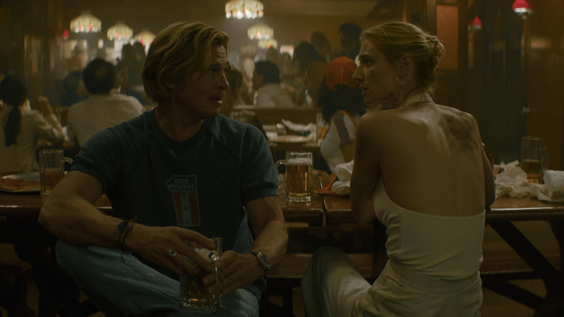 A man in a T-shirt and a woman in a white halter dress talk at a crowded bar, in a still from The Further Mis-Adventures of Cliff Booth