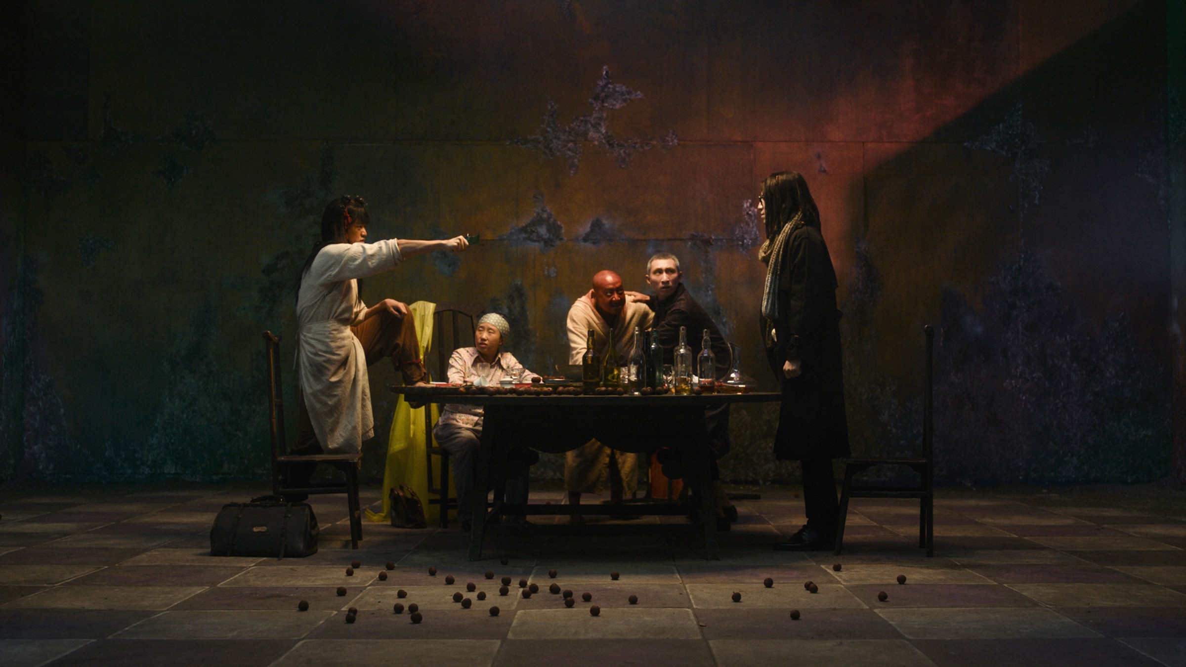 Diners seated at a long table on a painted stage set as a woman points across the room, in a scene from Fuxi: Four Tales of Feasting