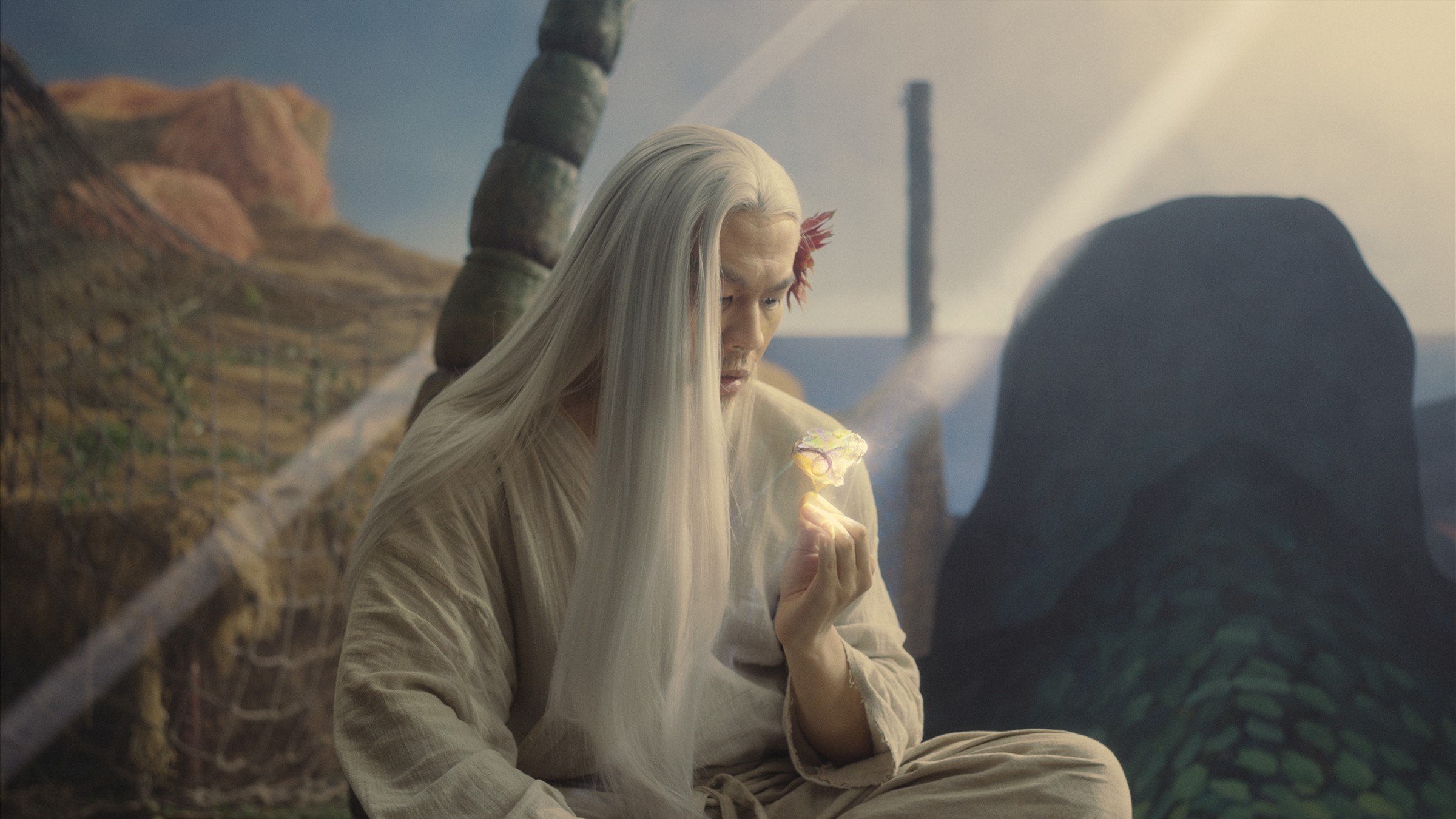 A white-haired figure in pale robes sits on a painted rocky set, holding a small glowing object, in a still from Fuxi: Four Tales of Feasting