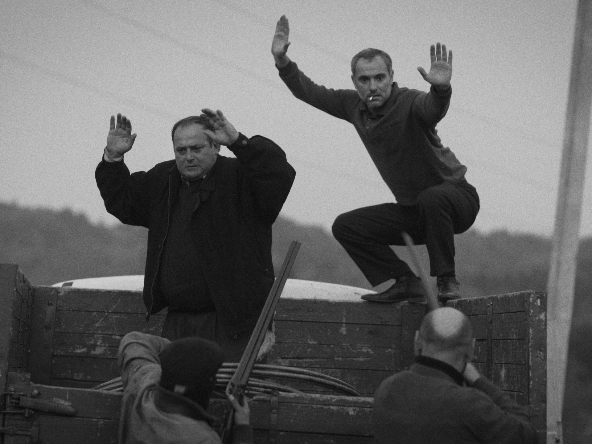 Black-and-white still of two men with their hands raised on the back of a truck, held up by men with a shotgun