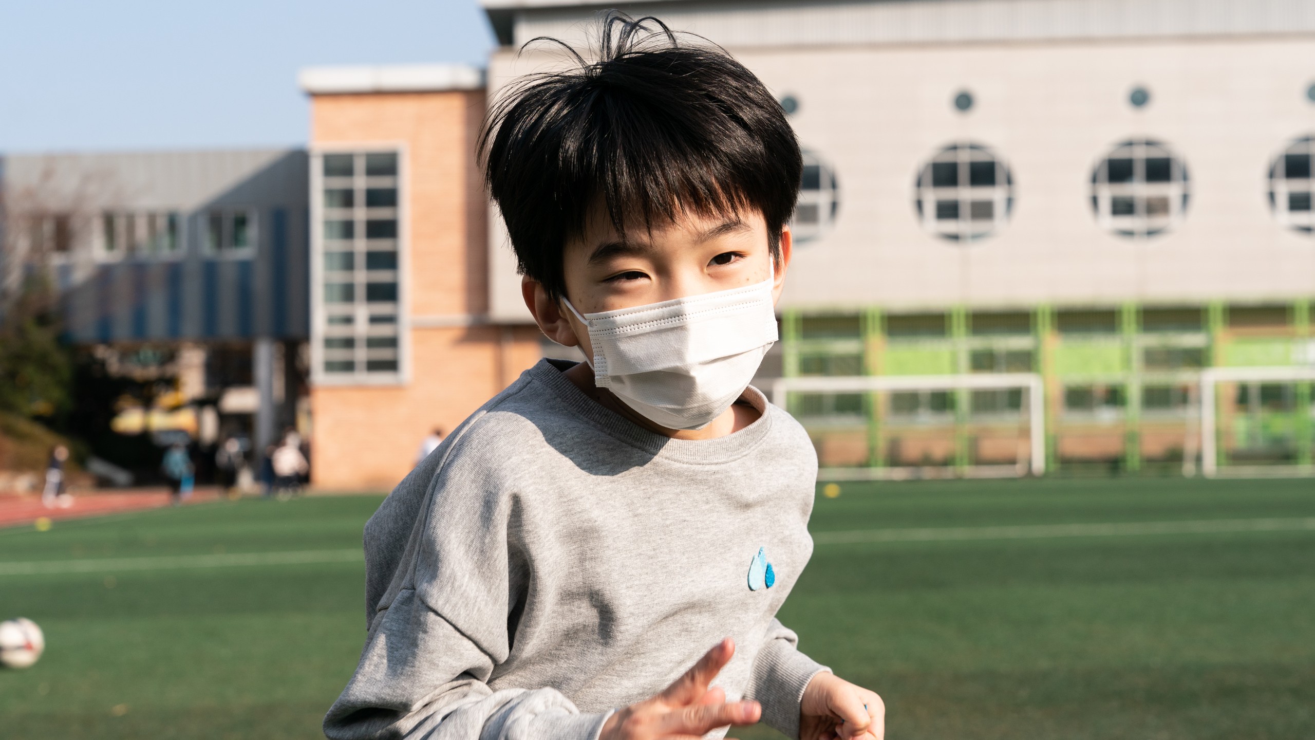 A boy in a face mask and grey sweatshirt runs across a school sports field in a still from Heartbeat