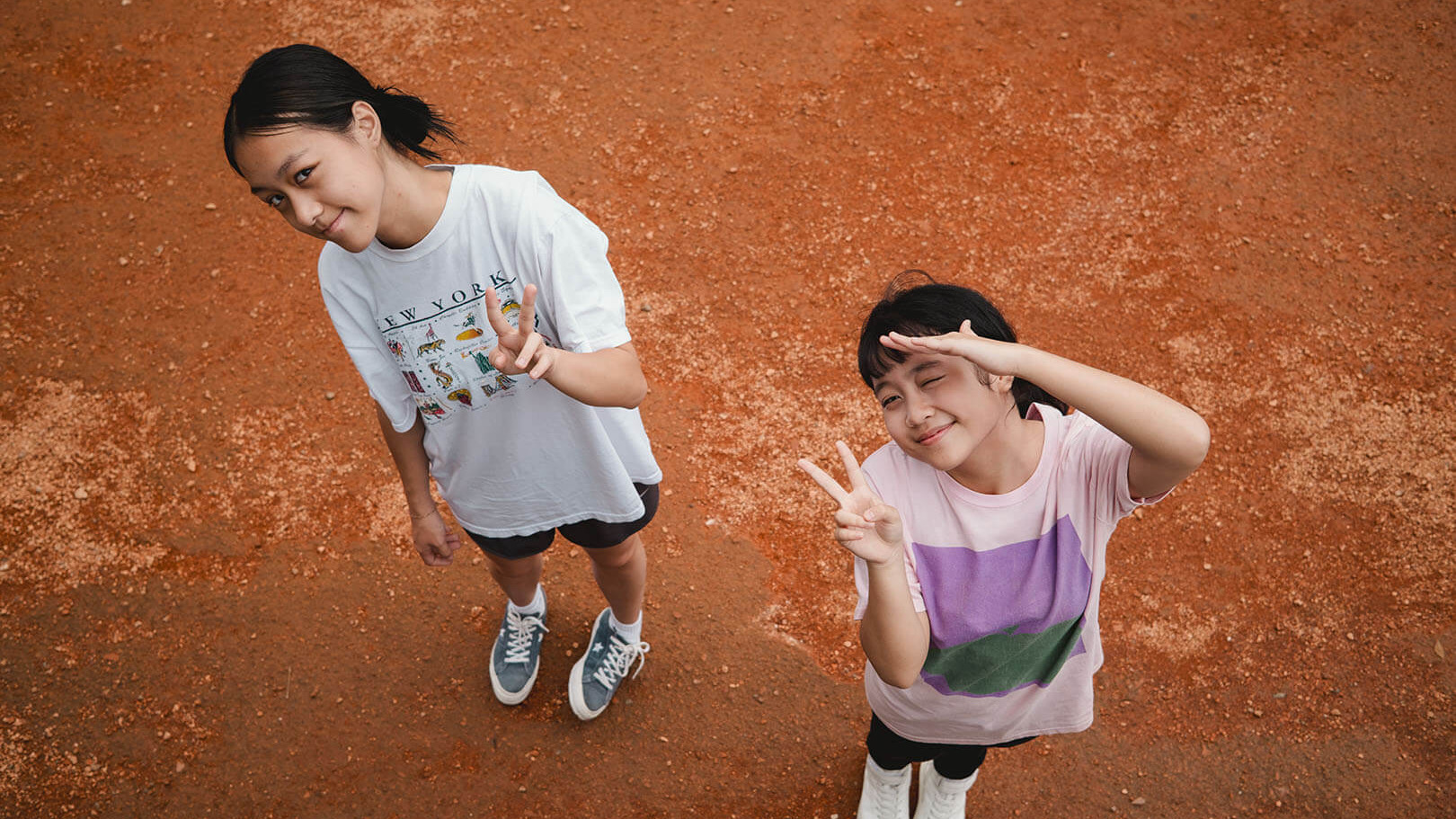 Two teenage girls in T-shirts, seen from above on red earth, smiling up at the camera and making peace signs