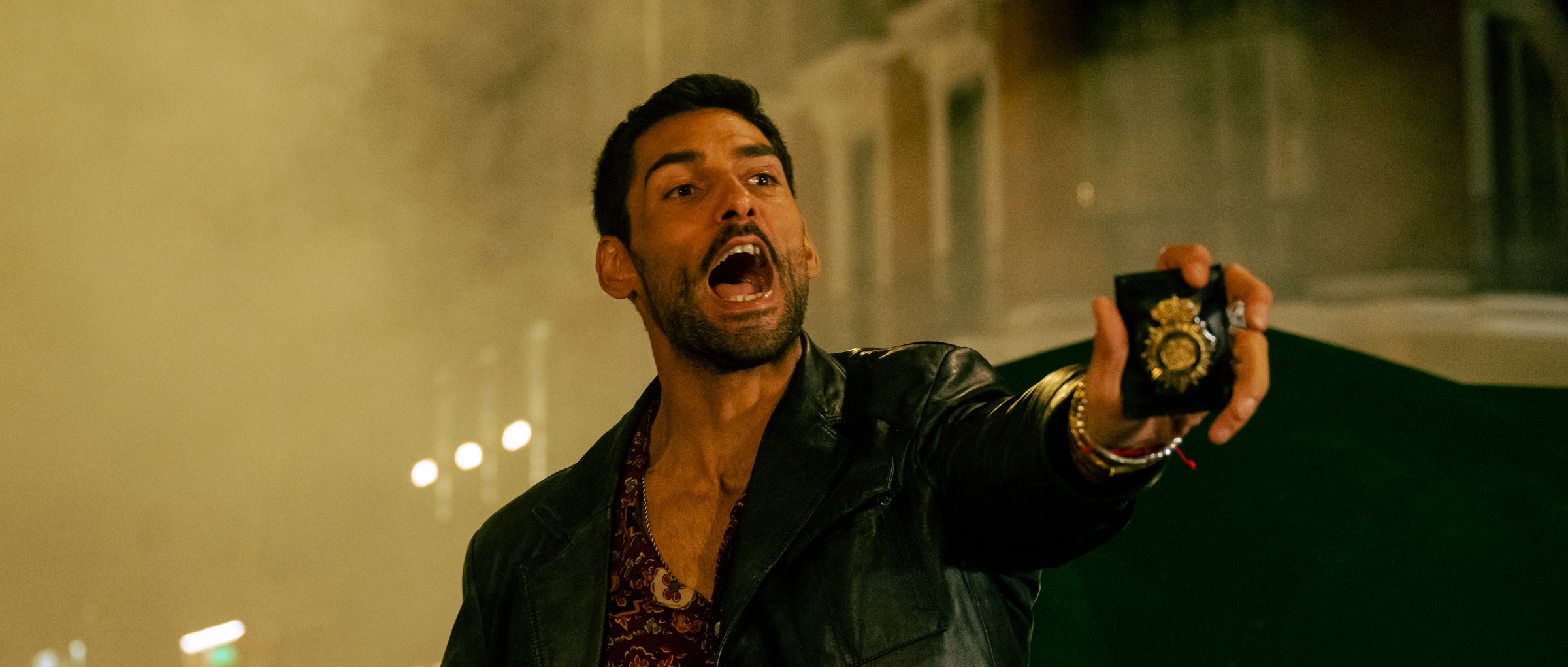 A man in a leather jacket shouts and holds up a police badge in a smoky street at night, in a still from Jotadé