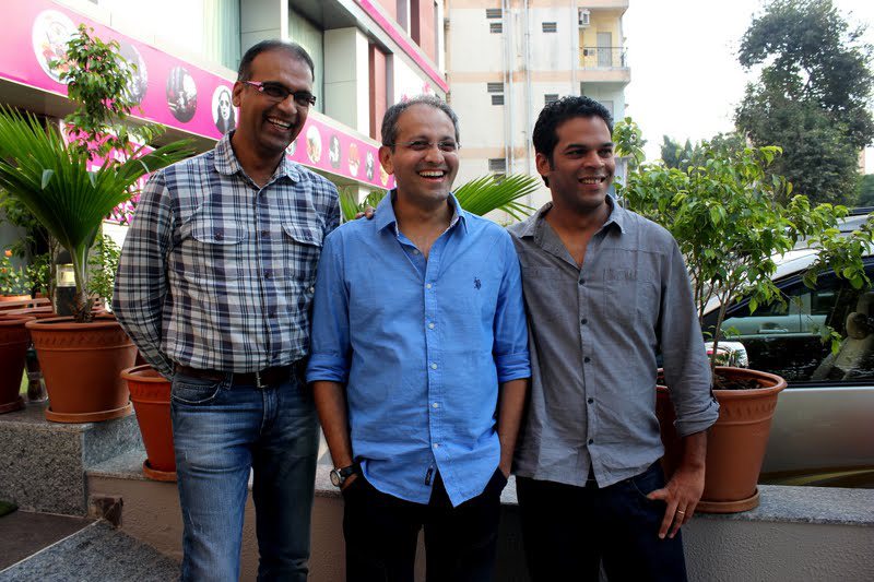 Three smiling men in checked, blue and grey shirts stand side by side beside potted plants outside a building