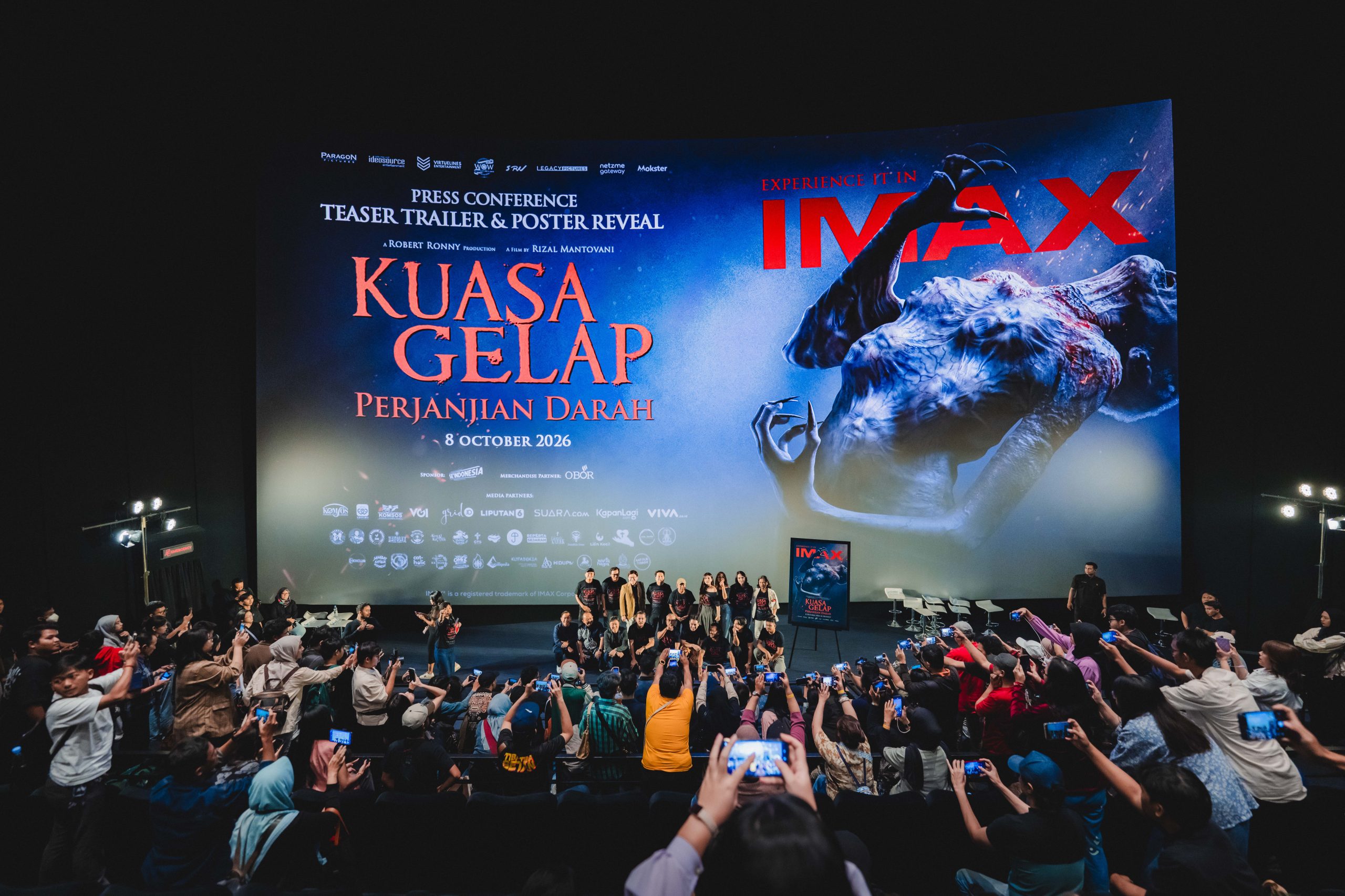Cast and crew line up on stage before a screen showing the Kuasa Gelap Perjanjian Darah title and IMAX logo as guests film on phones
