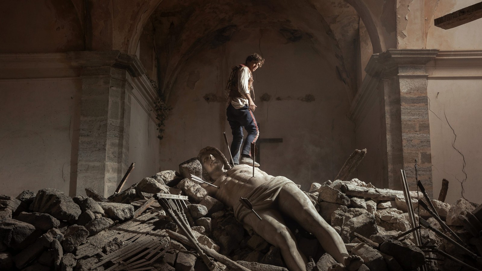 A young man stands on a pile of rubble and a fallen statue inside a ruined church, in a still from La bola negra