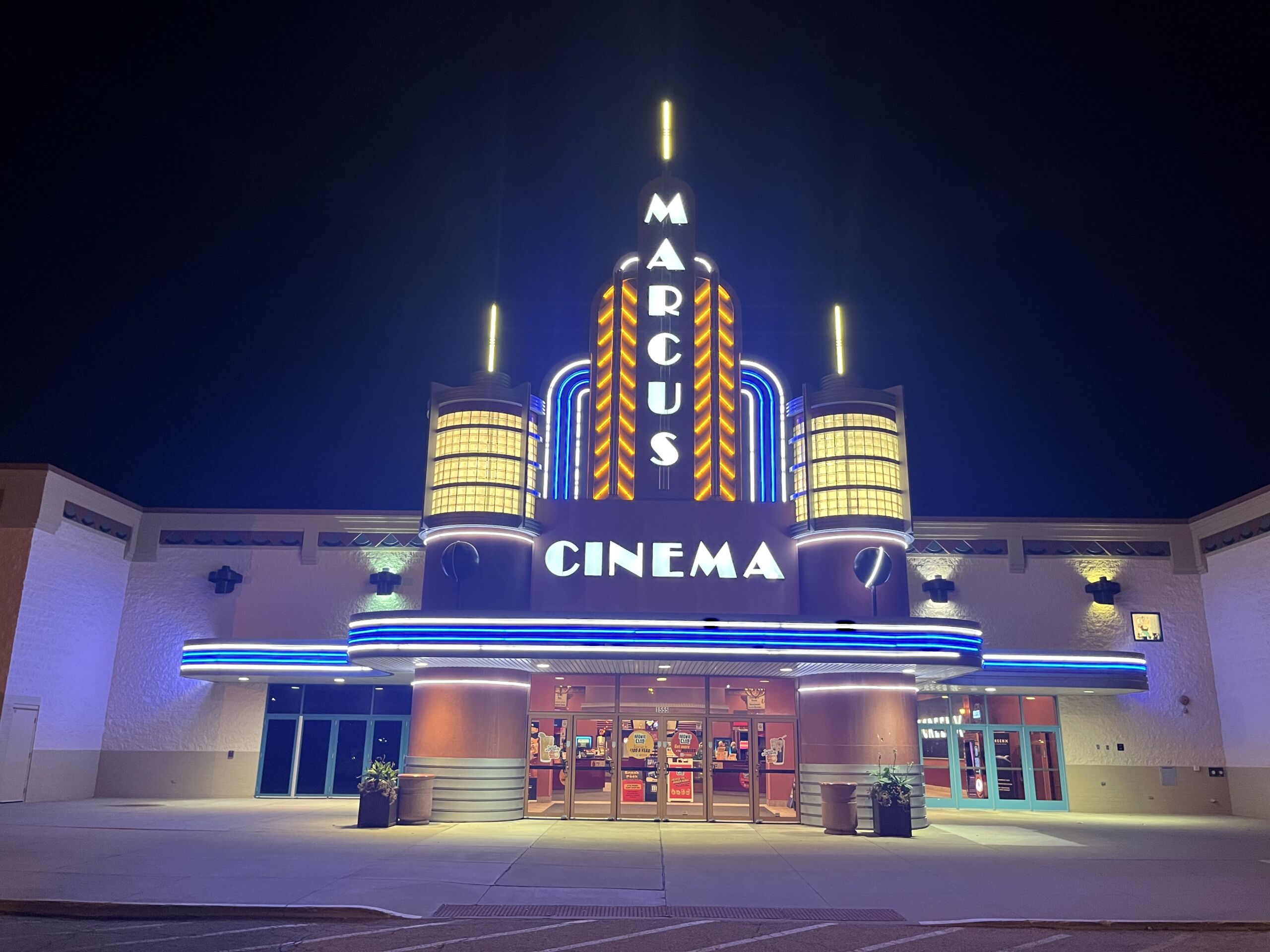 The front of the Marcus Addison Cinema at night, with a lit vertical Marcus sign, blue neon bands and Cinema lettering above glass doors
