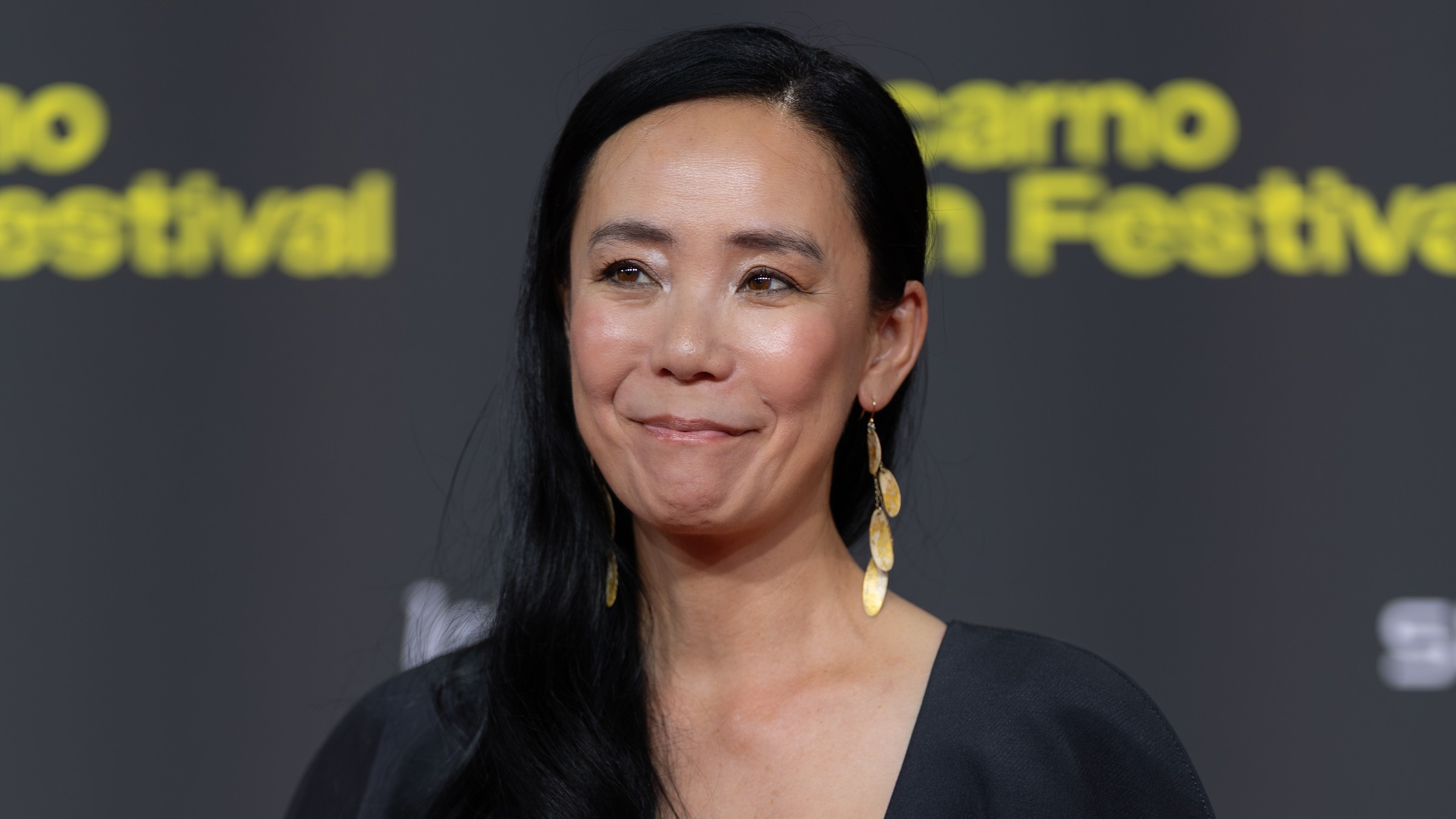 Naomi Kawase, with long dark hair and gold earrings, smiling on a red carpet in front of a Locarno Film Festival backdrop