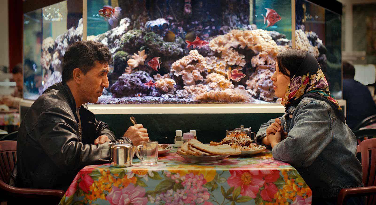 A man in a leather jacket and a woman in a floral headscarf talk at a restaurant table, in a still from No Good Men