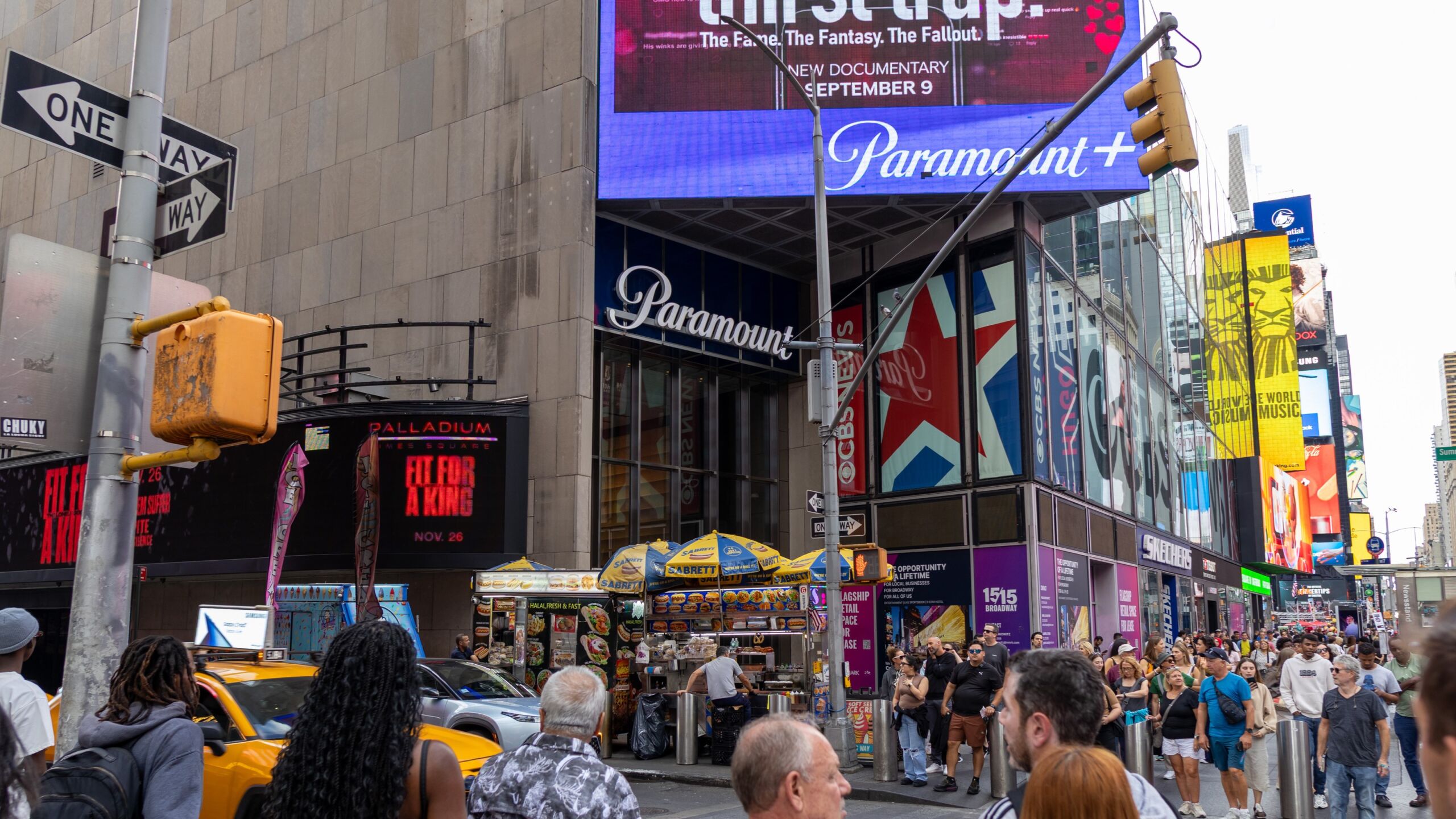 Pedestrians cross a Times Square street below a building with a Paramount sign and a Paramount+ video screen