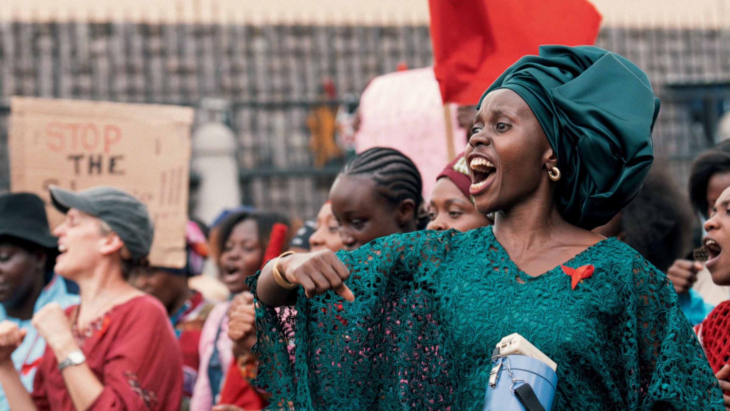 Eliane Umuhire, in a green headwrap and lace dress with a red ribbon, shouts and points at a street protest in Call Me Queen