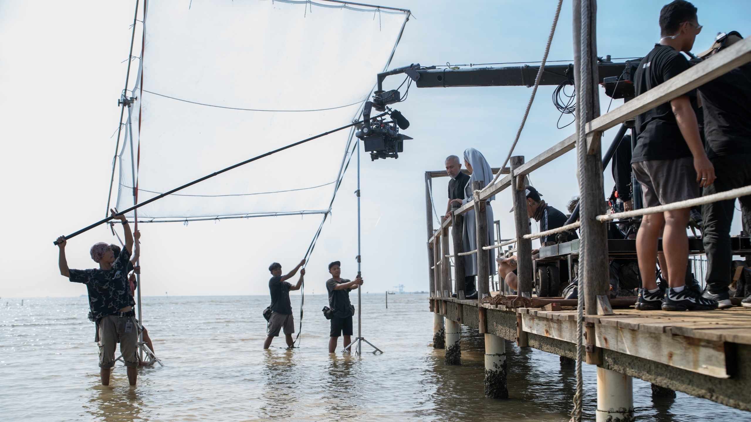 Crew members stand in shallow sea water holding a reflector as a priest and a nun in costume wait on a wooden pier by a camera crane