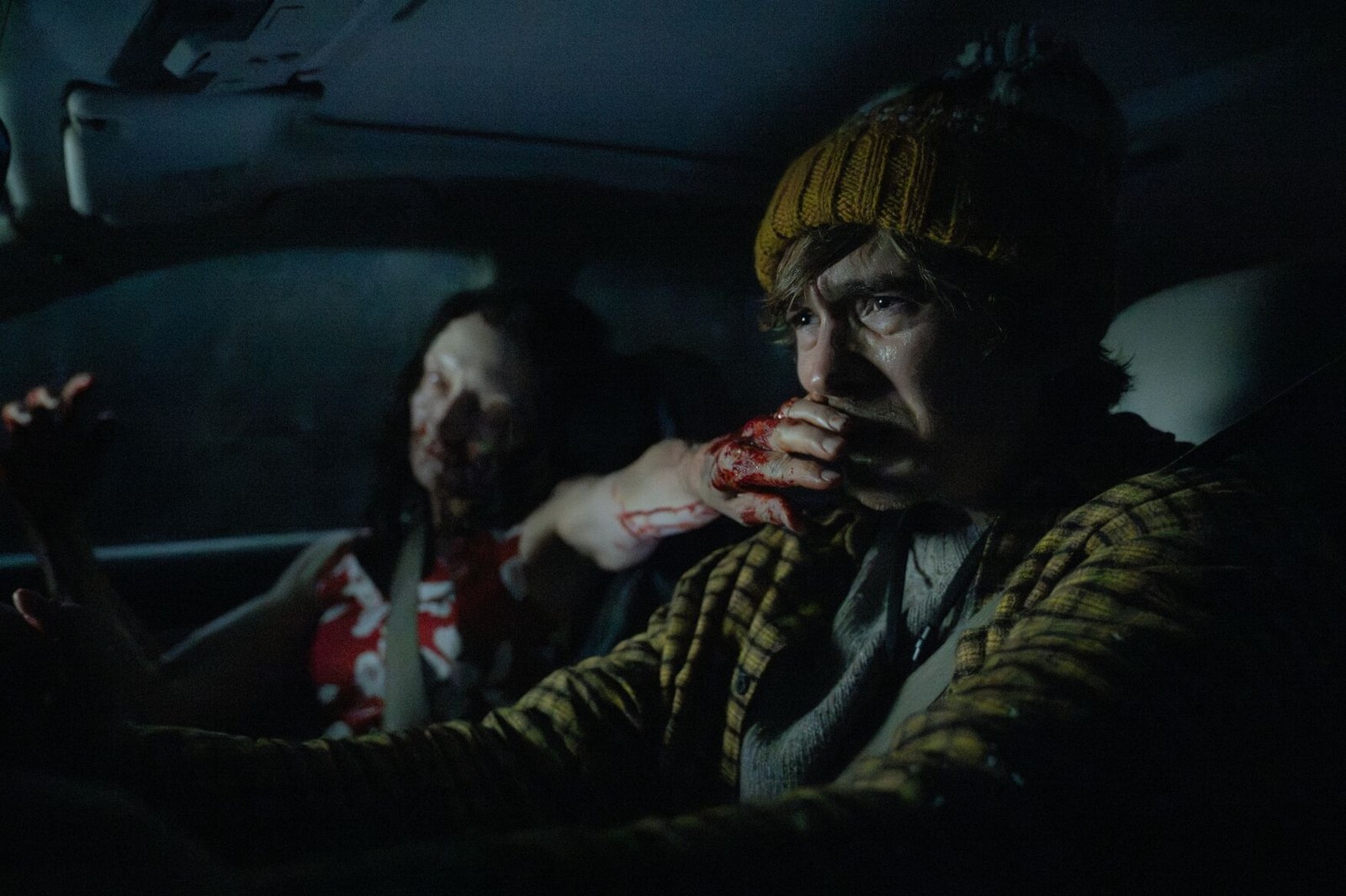 A frightened young man in a knitted hat drives at night as a bloodied hand clamps over his mouth from the passenger seat, in a still from Resident Evil