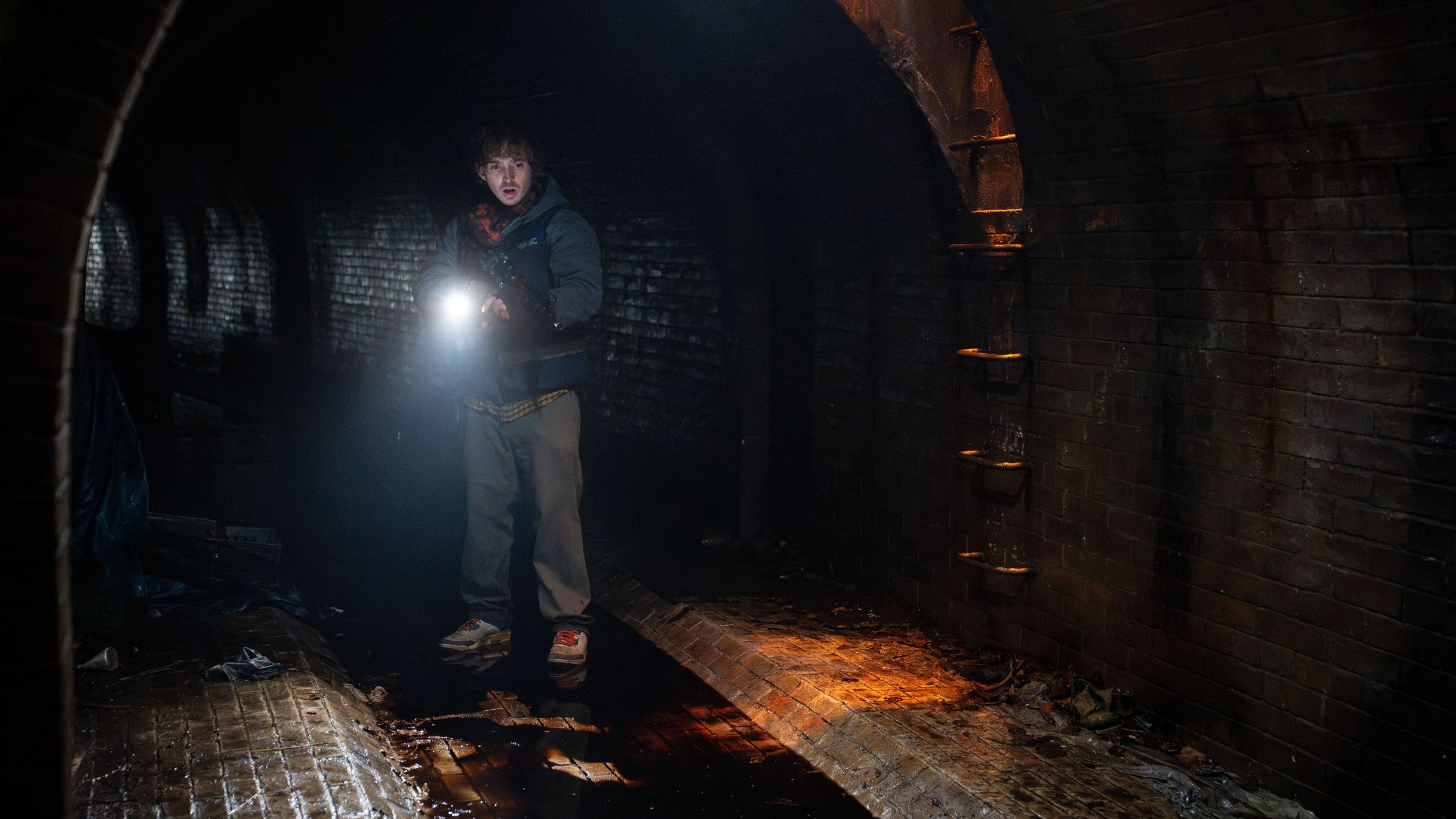 A young man in a padded jacket holds a torch in a dark brick tunnel with water on the floor, in a still from Resident Evil