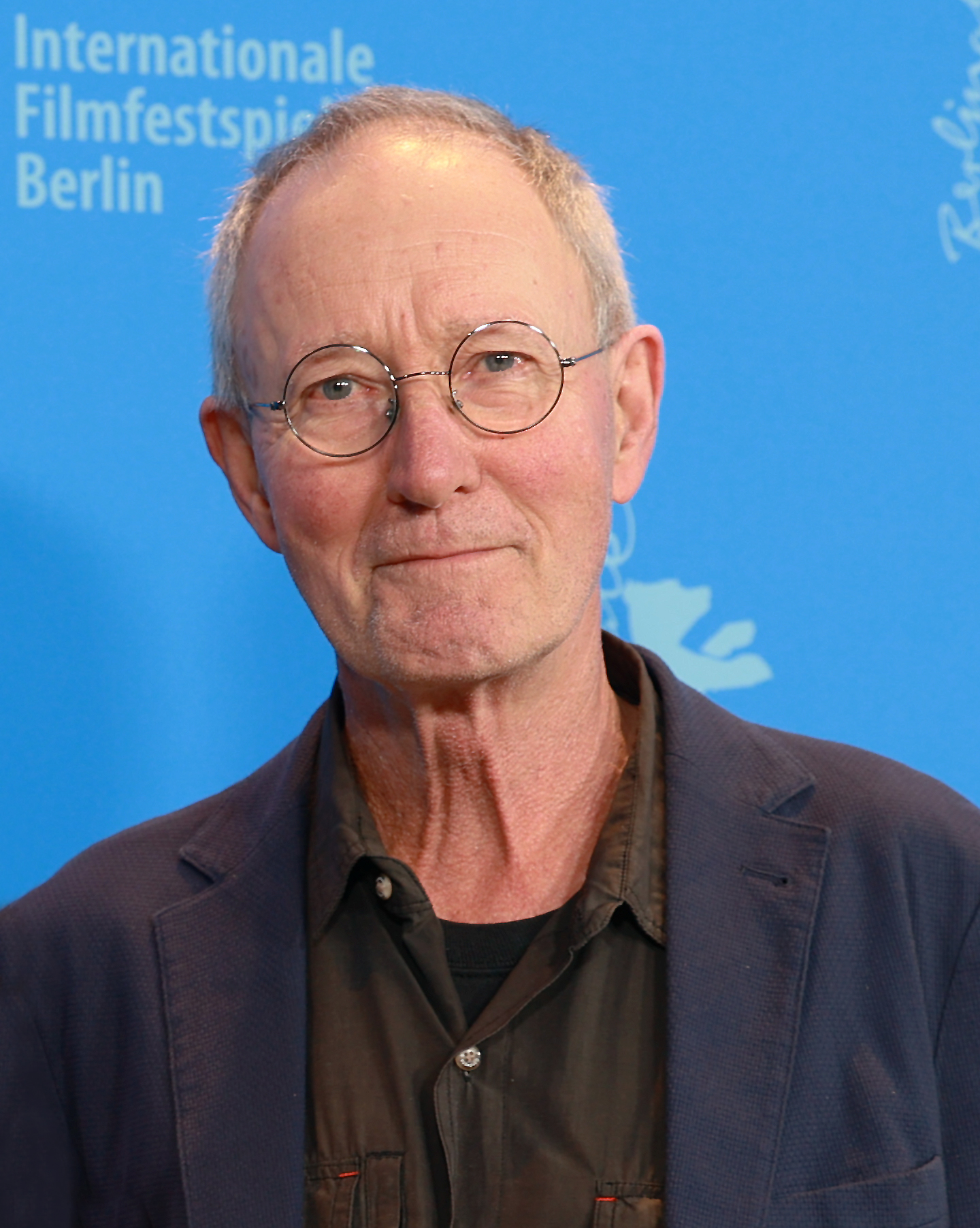 Rolf de Heer, white-haired with round wire-rimmed glasses, a dark jacket and an olive shirt, in front of a blue Berlinale backdrop