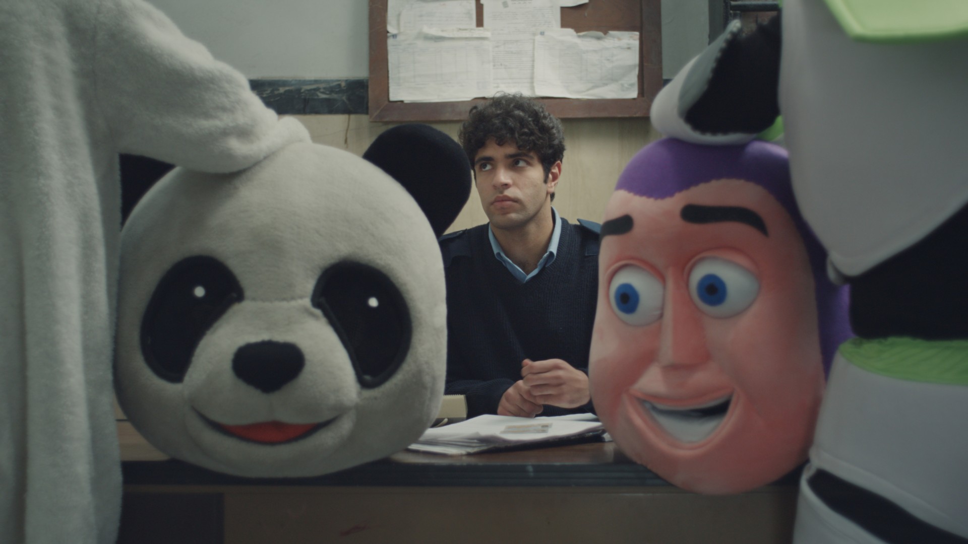 A young security guard sits at a desk behind two large mascot costume heads, a panda and a cartoon face