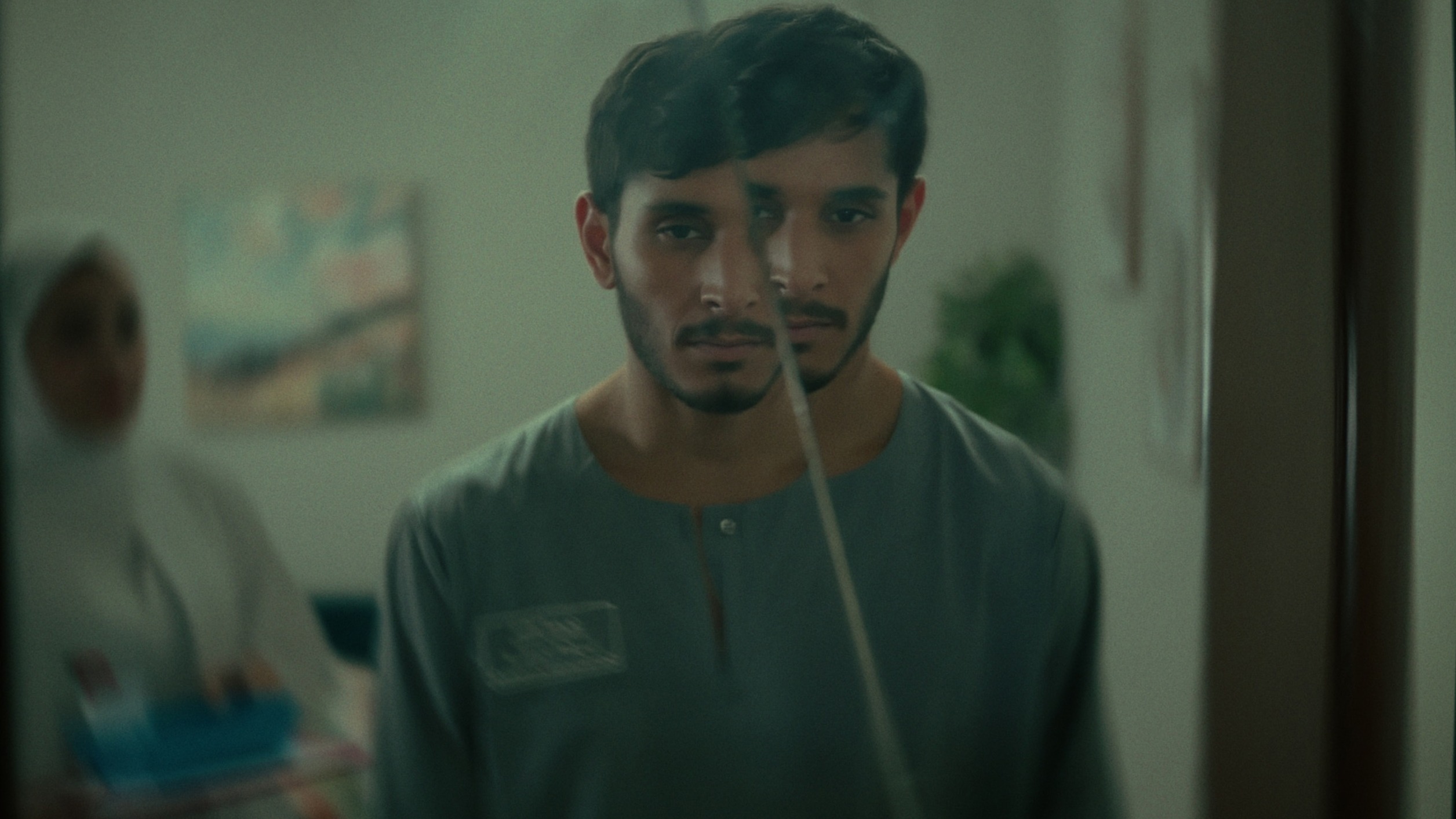 A young man in a grey-green tunic behind a cracked pane of glass, a woman in a white headscarf behind him, in Salem Not Salem