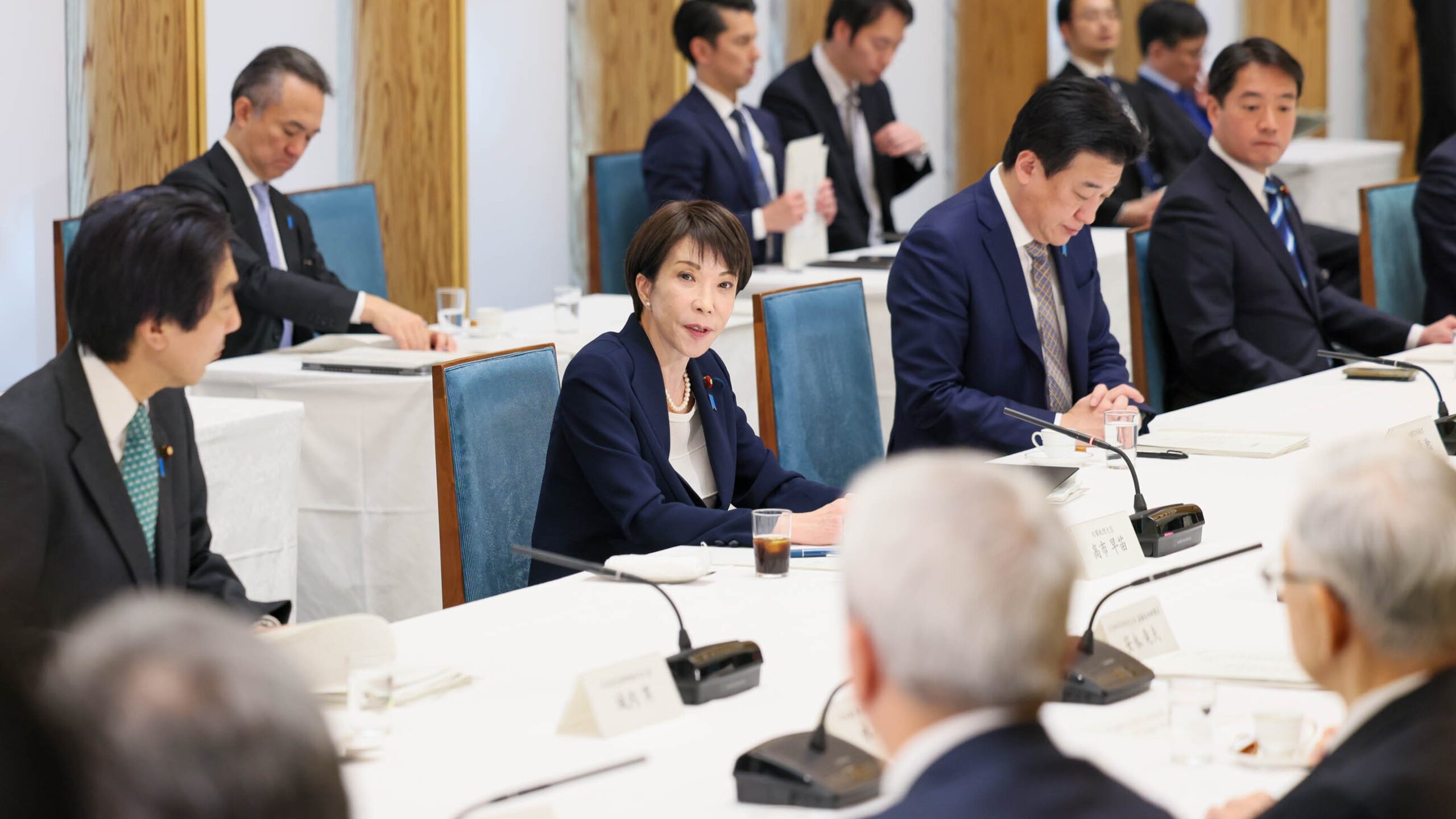 Sanae Takaichi, in a navy jacket, speaks at a long conference table among suited business leaders and officials