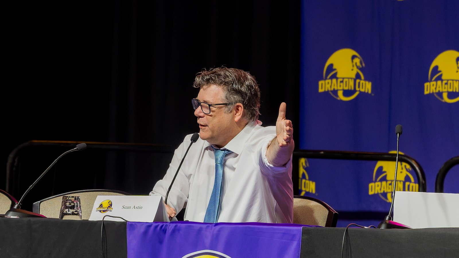 Sean Astin in glasses, a white shirt and blue tie, gesturing with one hand at a panel table with a microphone and name card