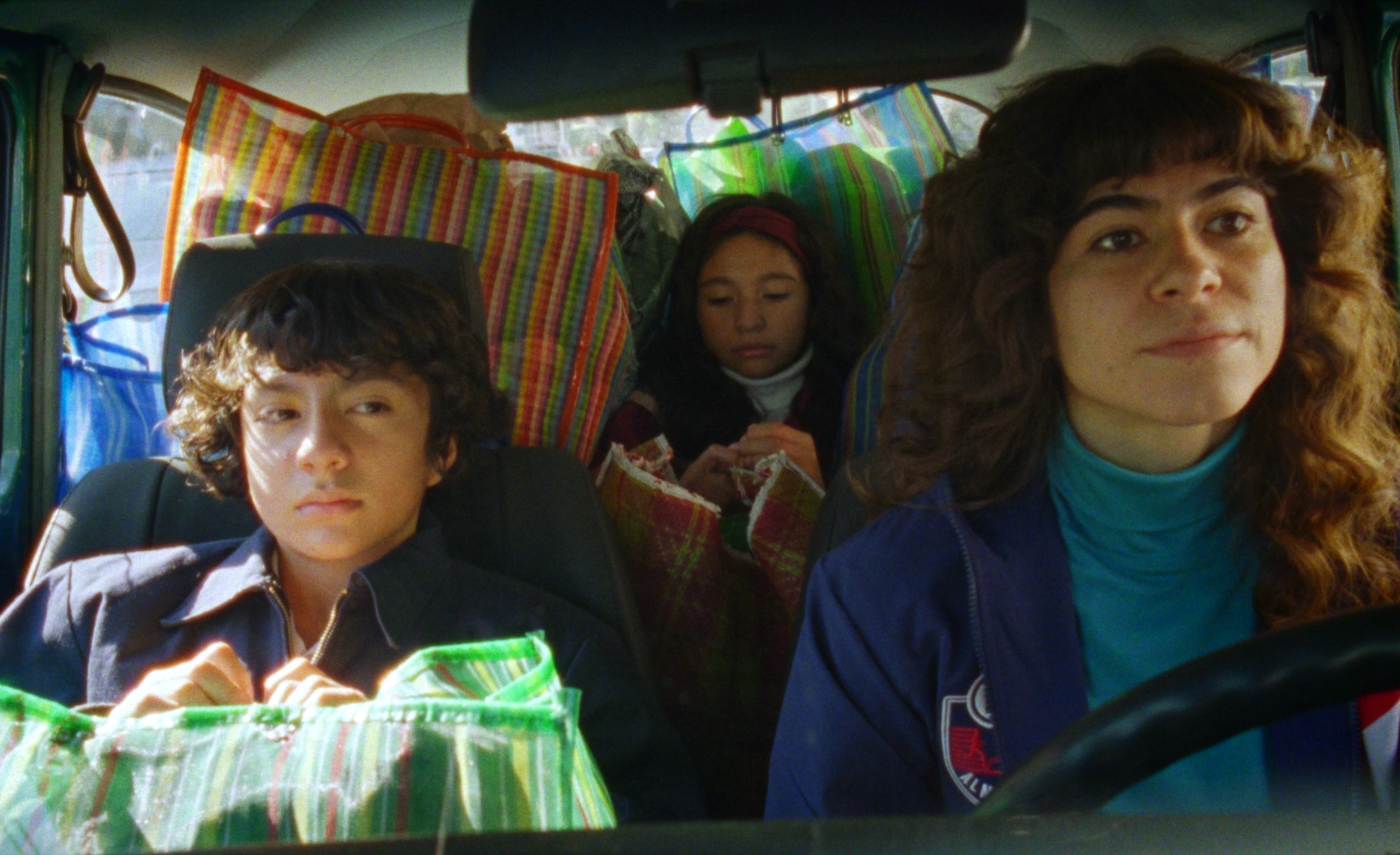 A boy holding a striped bag sits beside a curly-haired woman driving a car, with a girl and more bags in the back seat