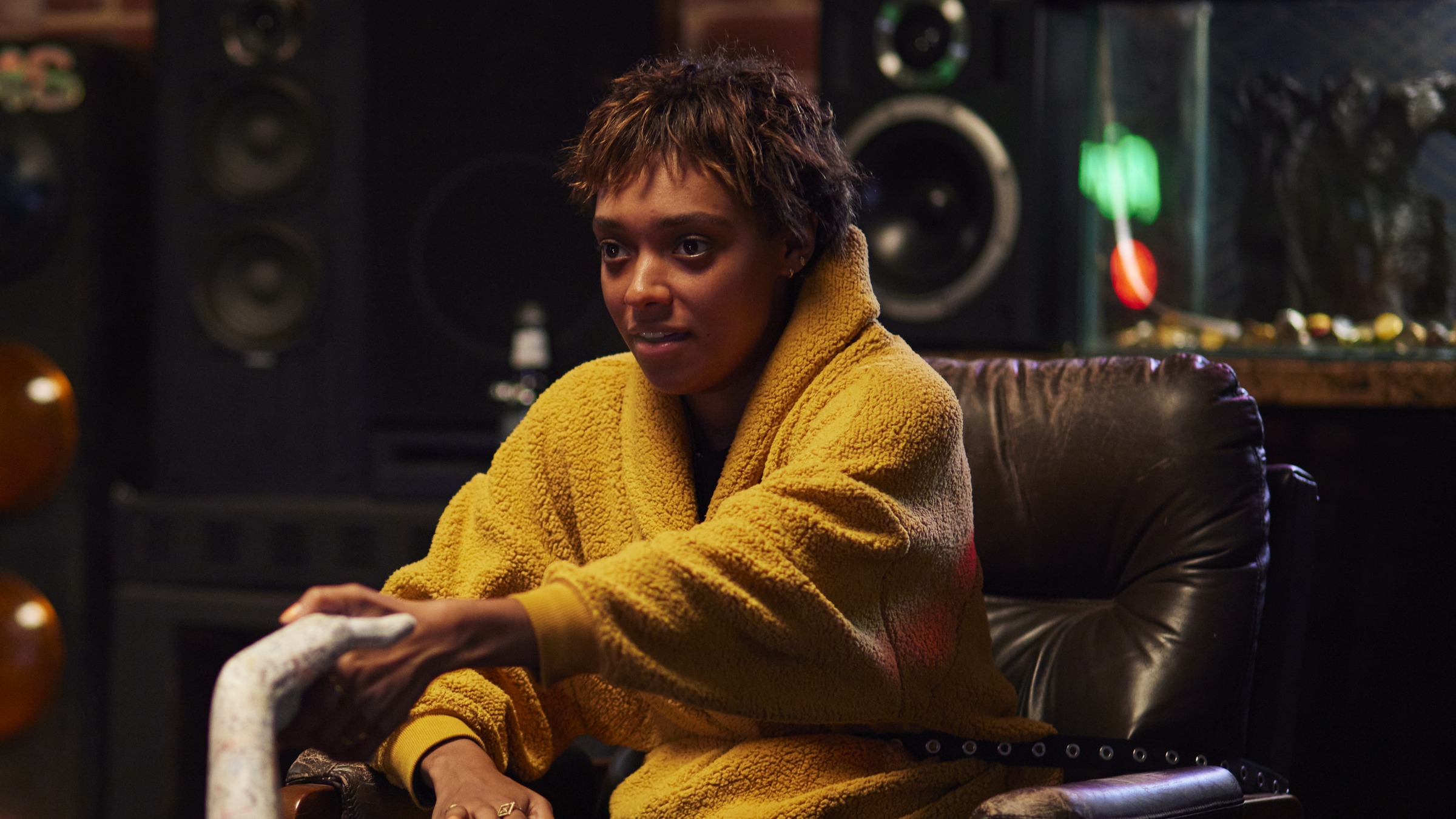 A young woman in a mustard fleece jacket sits in a leather armchair with her hand on a white sculpted hand, in a dim room lined with speakers, in a still from Talk To Me