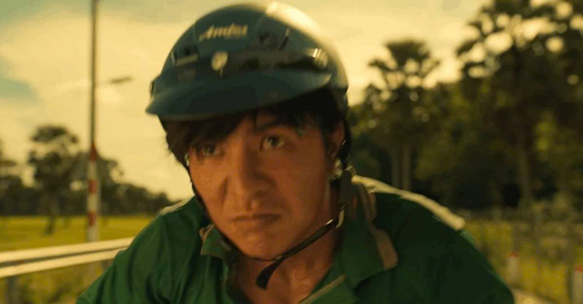 A young man in a green shirt and dark helmet stares ahead on a rural road, in a still from Trại buôn người