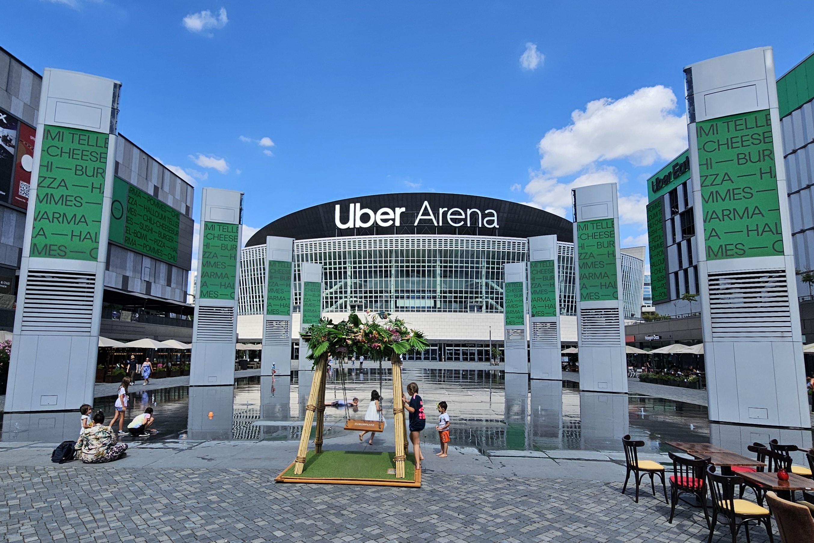 The curved glass front of the Uber Arena in Berlin under a blue sky, seen across Uber Platz