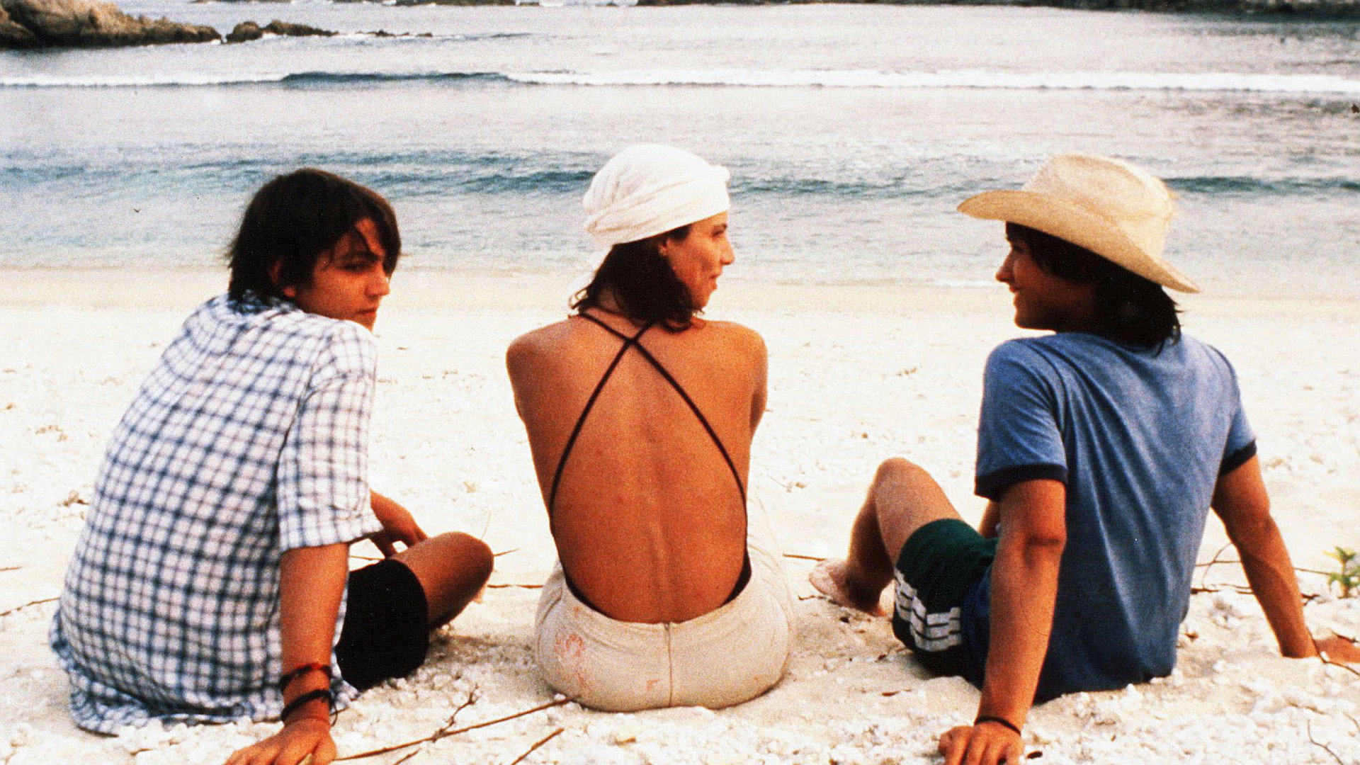 Two young men and a woman in a white headscarf sit on a white-sand beach facing the sea, in a still from Y tu mamá también